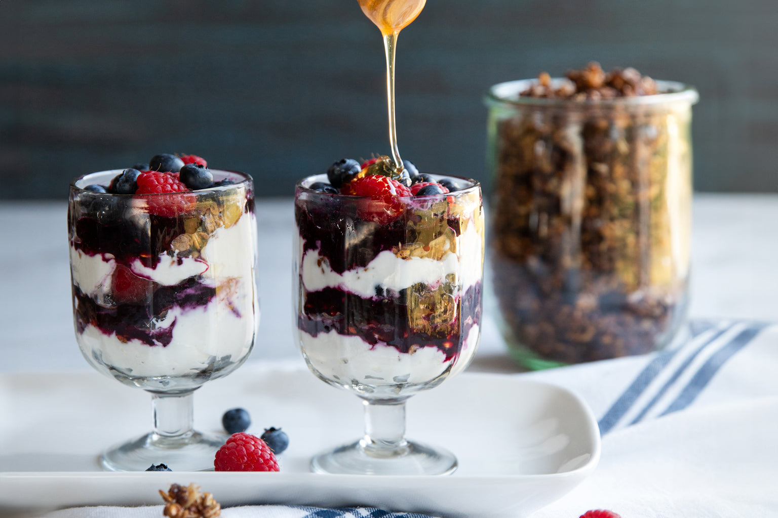Two glasses filled with berry and yogurt parfaits with einkorn wheat berry granola, one being drizzled with honeyeinkorn wheat berry granola parfait