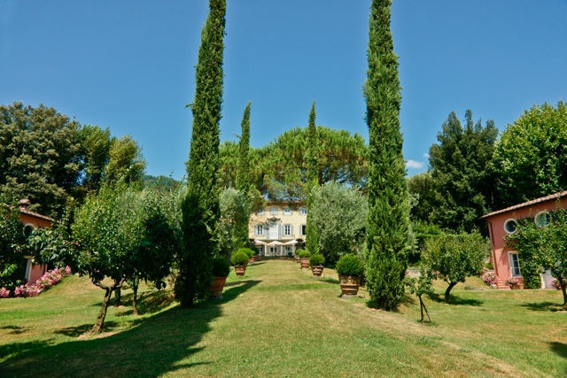 Cream-colored villa at end of a cypress-lined lawn with terracotta potted shrubs and pink outbuildings under a blue sky