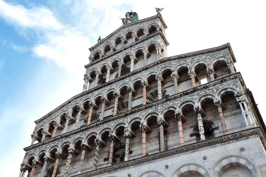 Romanesque cathedral facade with stacked arched loggias, patterned columns and an angel statue at the top against a blue sky.