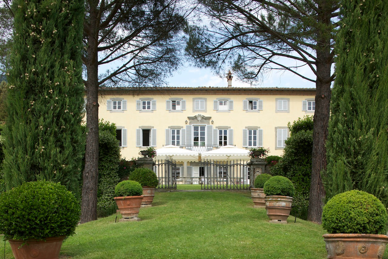 Yellow villa with shuttered windows, terracotta planters and boxwood leading to a wrought-iron gate and terrace umbrellas.