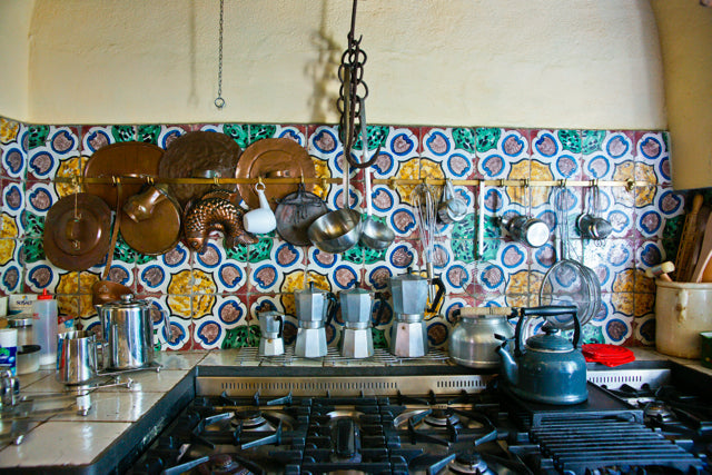 Italian style stove and gas burners with colorful patterned tile backsplash, hanging copper pans, three moka pots, and a black kettle.