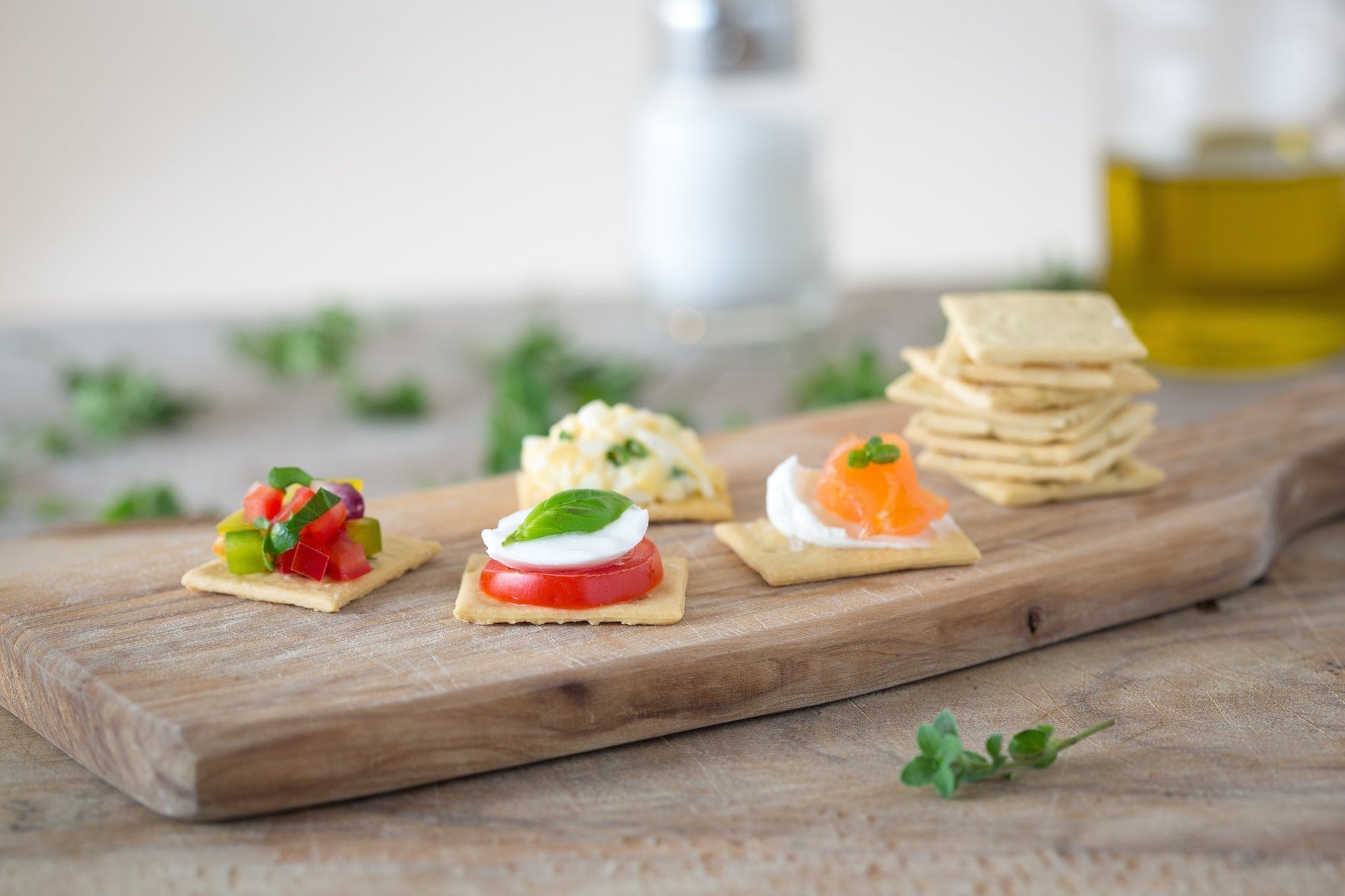 Crackers on a wooden board topped with tomato and mozzarella, smoked salmon, egg salad, and herb garnishes