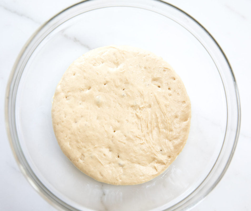 Round risen dough proofing in a glass mixing bowl on a marble countertop