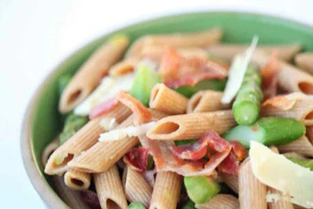 Bowl of penne with asparagus, crispy pancetta, and shaved Parmesan on a light background