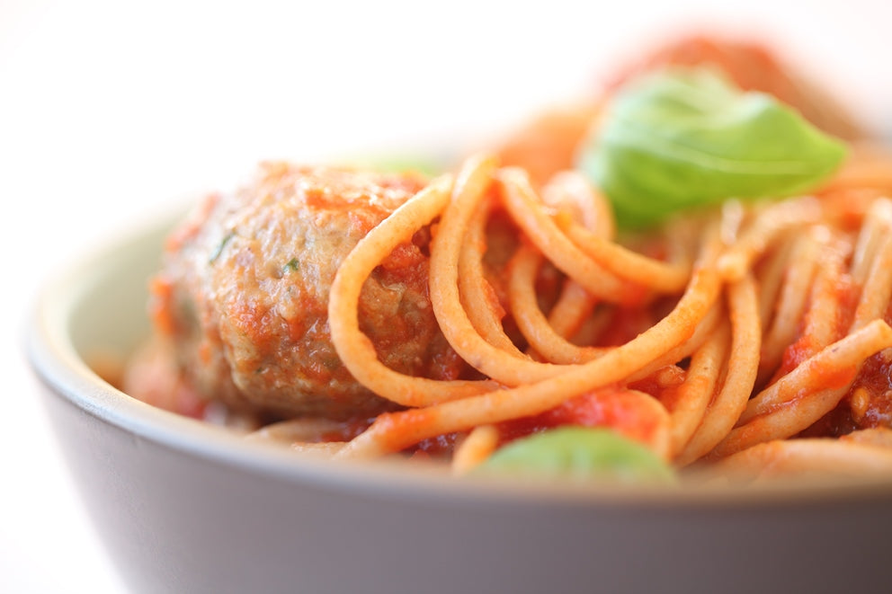 Close-up of spaghetti twirled with tomato sauce and a wheat-free meatball in a shallow bowl, garnished with fresh basil