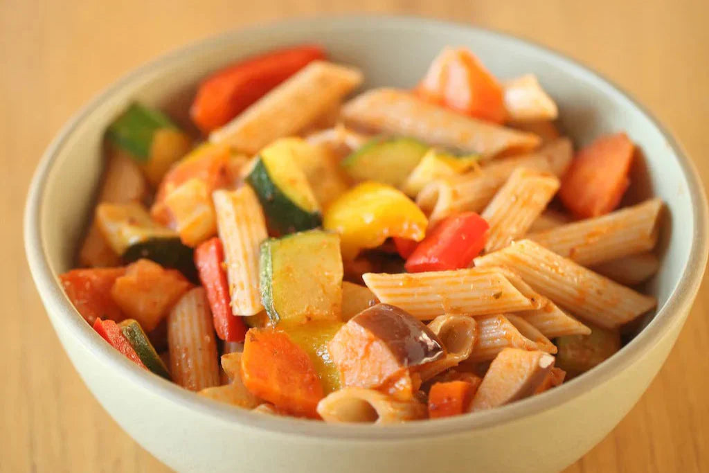 Bowl of penne with caponata of eggplant, zucchini, bell peppers and carrots on a wooden table