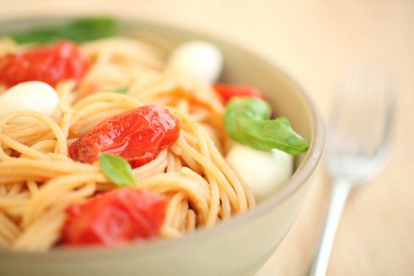 Bowl of gluten-free brown rice spaghetti with sautéed cherry tomatoes, ciliegine mozzarella and fresh basil, fork beside the bowl
