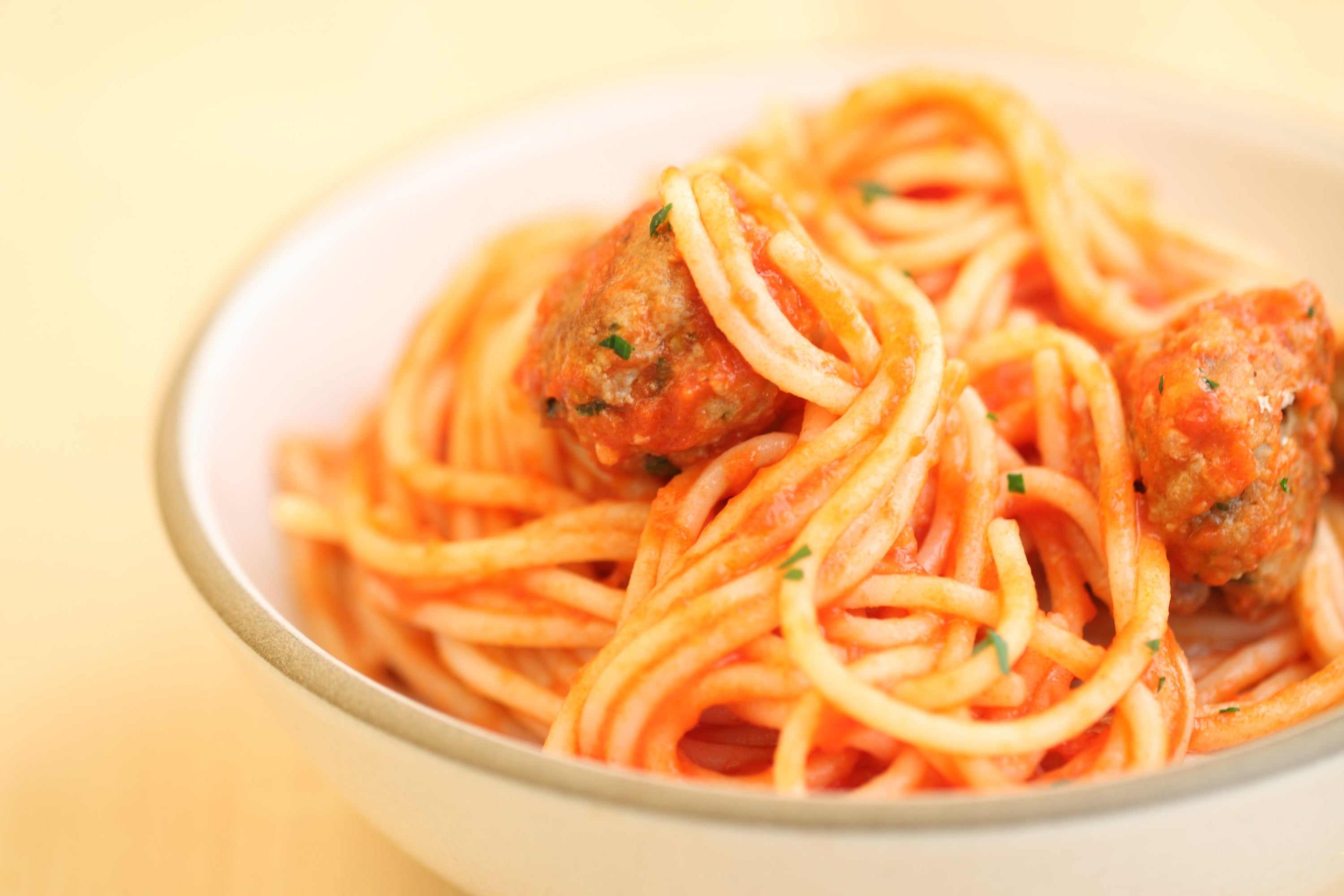 Bowl of spaghetti with Gluten Free meatballs in tomato sauce, garnished with parsley, on a pale background
