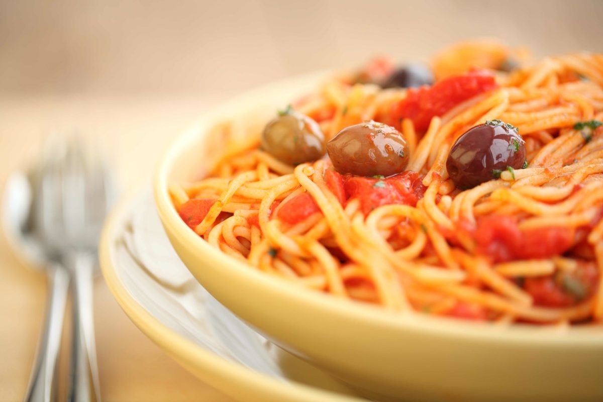 Bowl of spaghetti puttanesca with tomato sauce, olives, and basil in a yellow bowl with a blurred fork