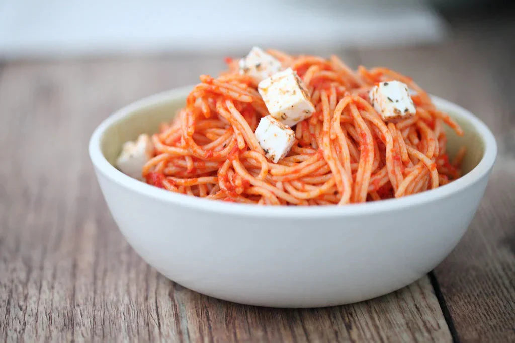 Bowl of gluten free brown rice spaghetti in red pepper and sun dried tomato sauce topped with feta cubes