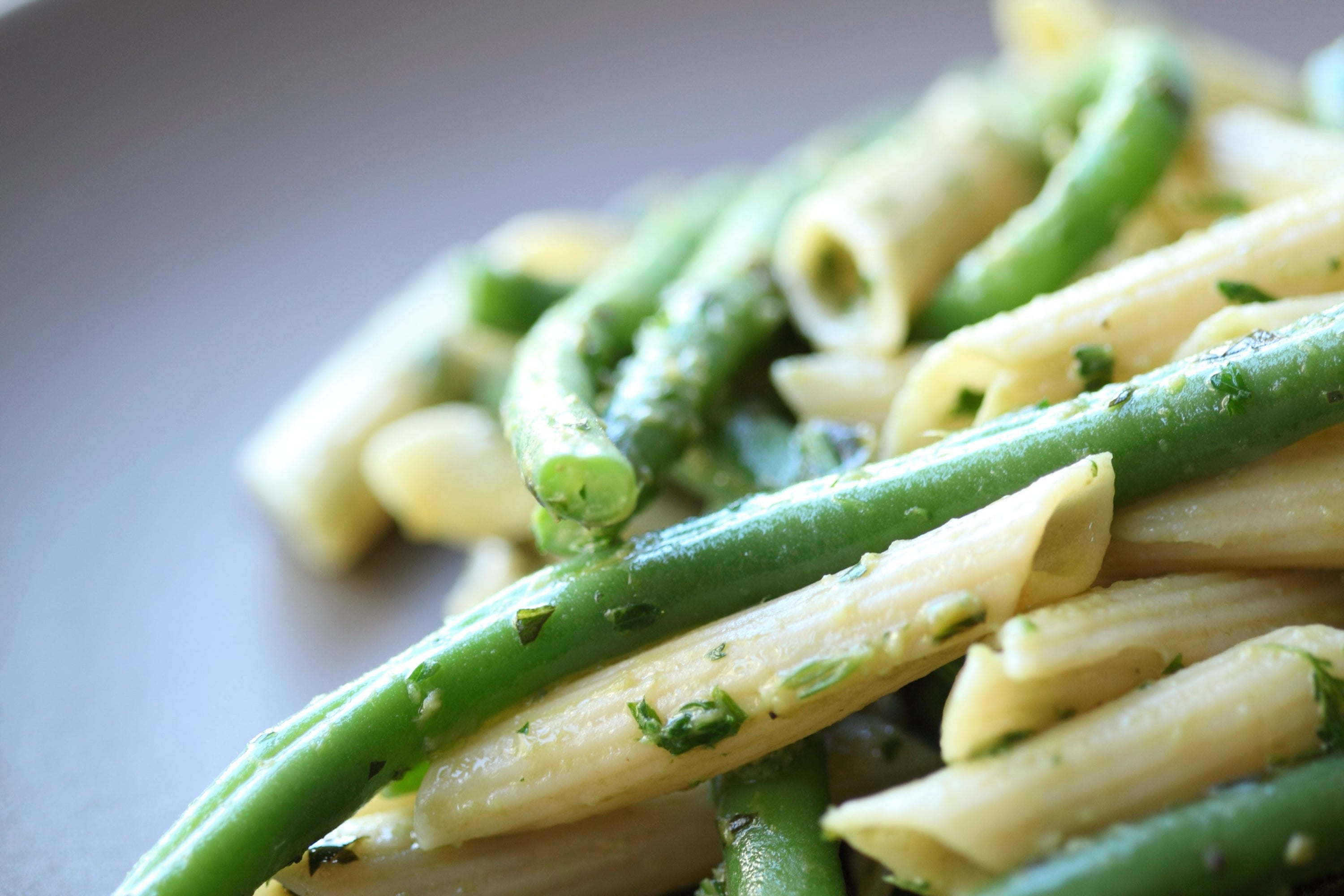Close up of brown rice penne tossed with green beans in a garlic, anchovy and herb sauce on a neutral background
