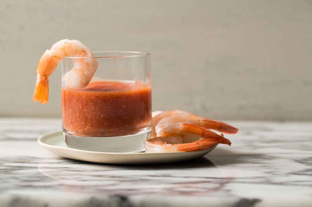Glass of Jovial cocktail sauce with cooked shrimp on a small plate, one shrimp hanging on the glass rim on a marble countertop