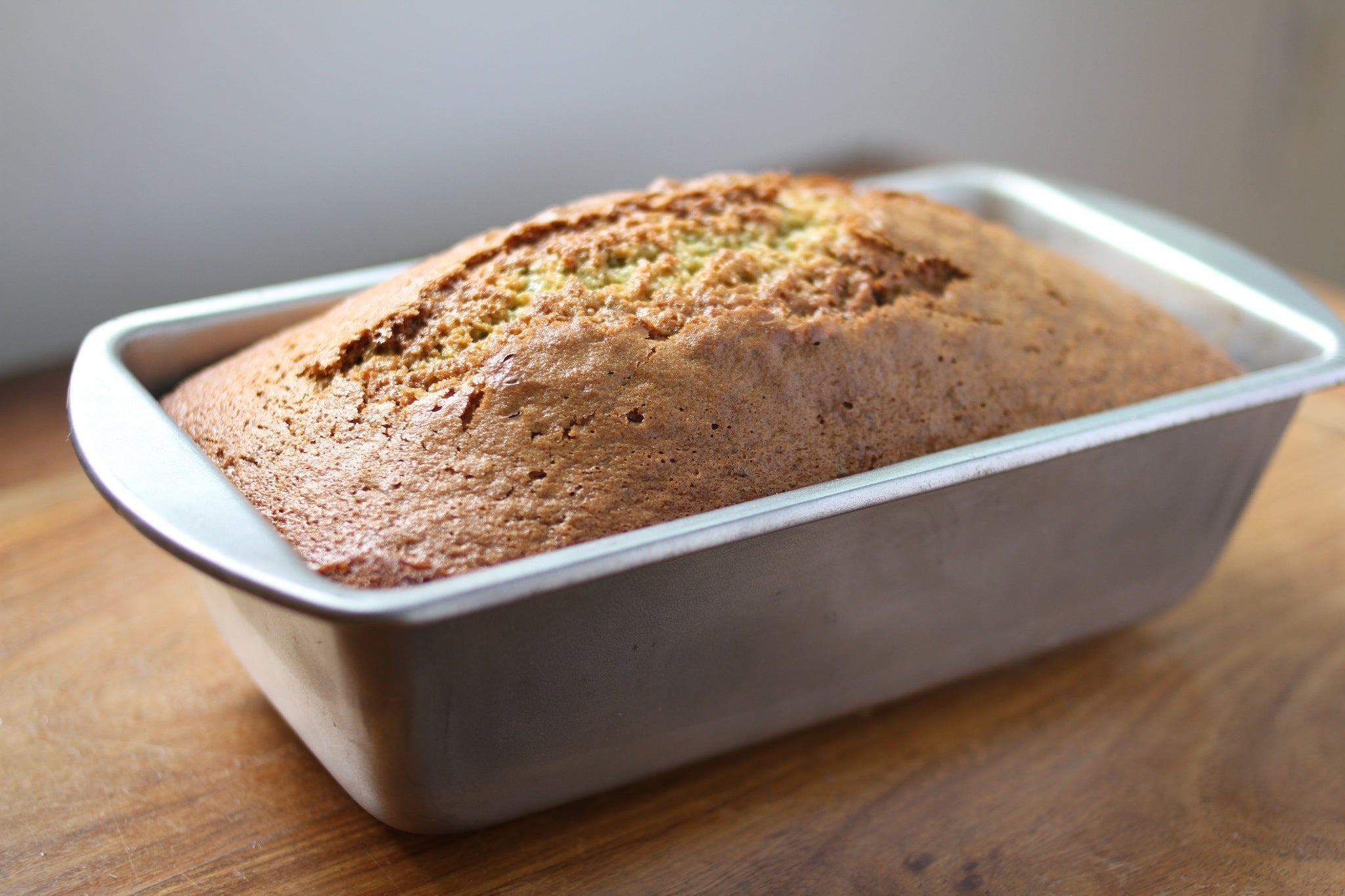 Three-quarter view of an Einkorn zucchini bread loaf in a metal loaf pan showing a golden-brown cracked top on a wooden countertop