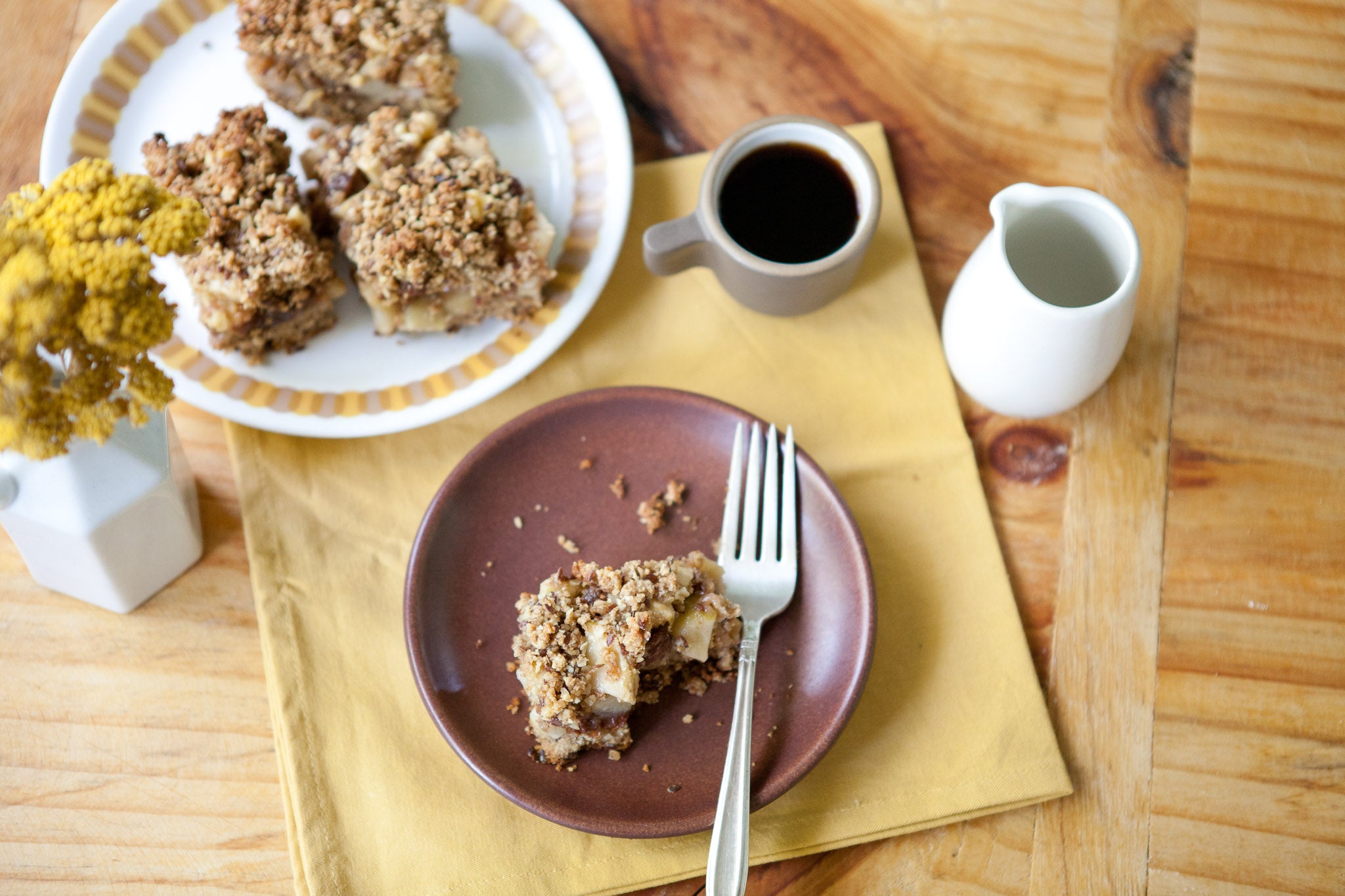 Apple raisin walnut squares served on a small brown plate with a fork, beside coffee and a milk jug on a wooden table
