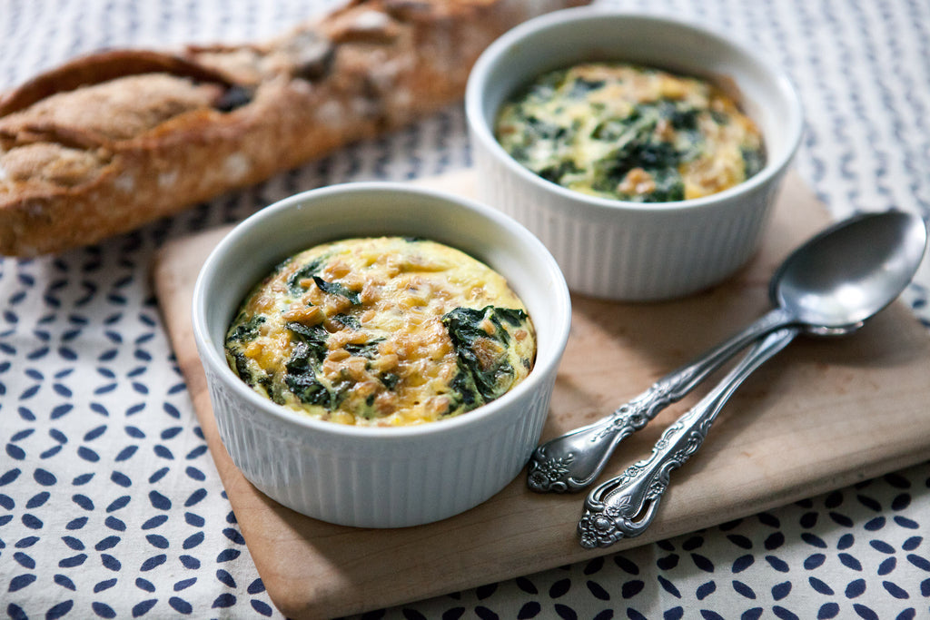 Two egg bakes with wheatberries, kale, and melted cheddar in white ramekins on a wooden board with a baguette and vintage spoons
