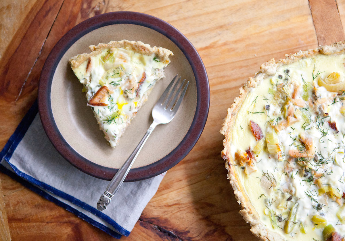 Slice of smoked salmon tart with an einkorn herbed crust on a ceramic plate with a fork, beside the remaining tart on a wooden table
