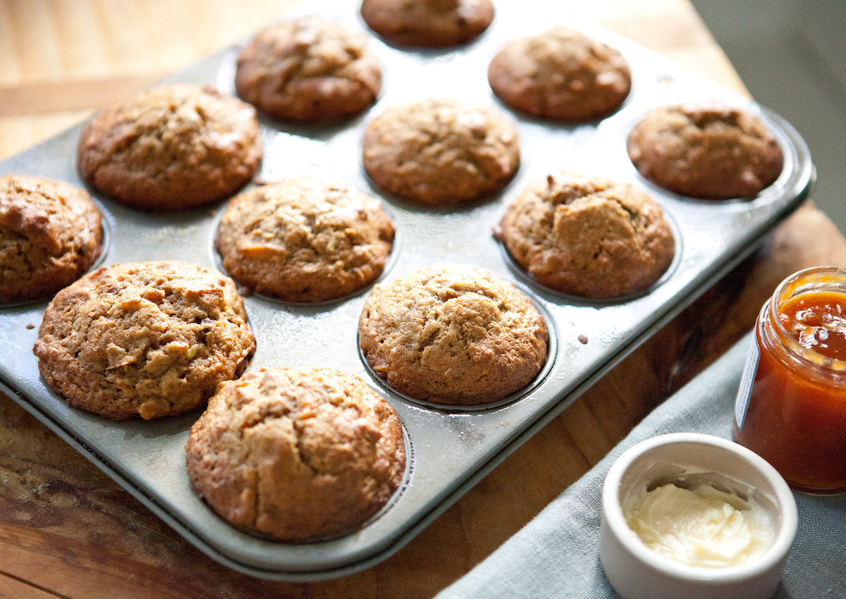 Freshly baked Einkorn morning glory muffins cooling in a muffin tin on a wooden board beside a jar of marmalade and a dish of butter