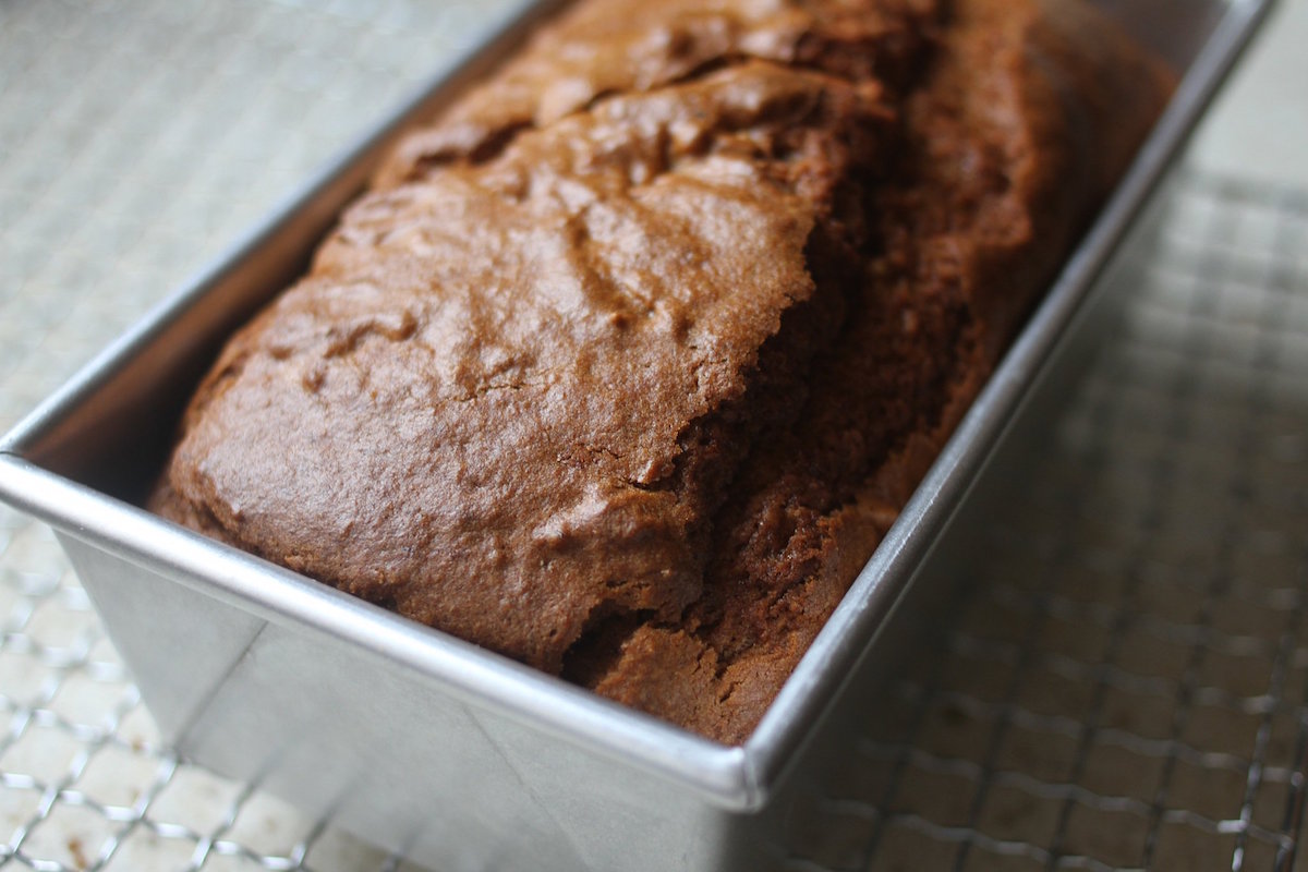 Close-up of whole-grain Einkorn pumpkin bread in a metal loaf pan, cracked golden-brown top and moist crumb on a cooling rack