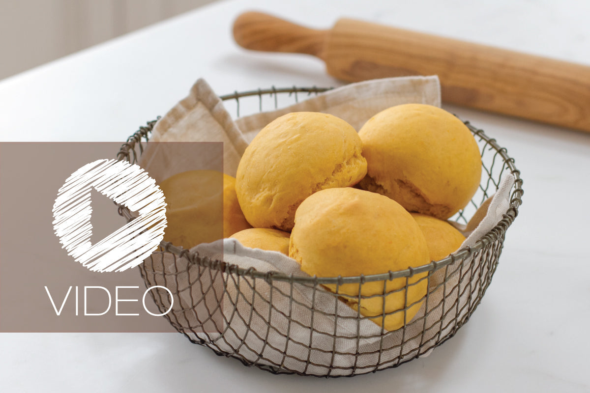 Front view of Einkorn sweet potato rolls in a wire basket with video play overlay, golden rounds on a white counter