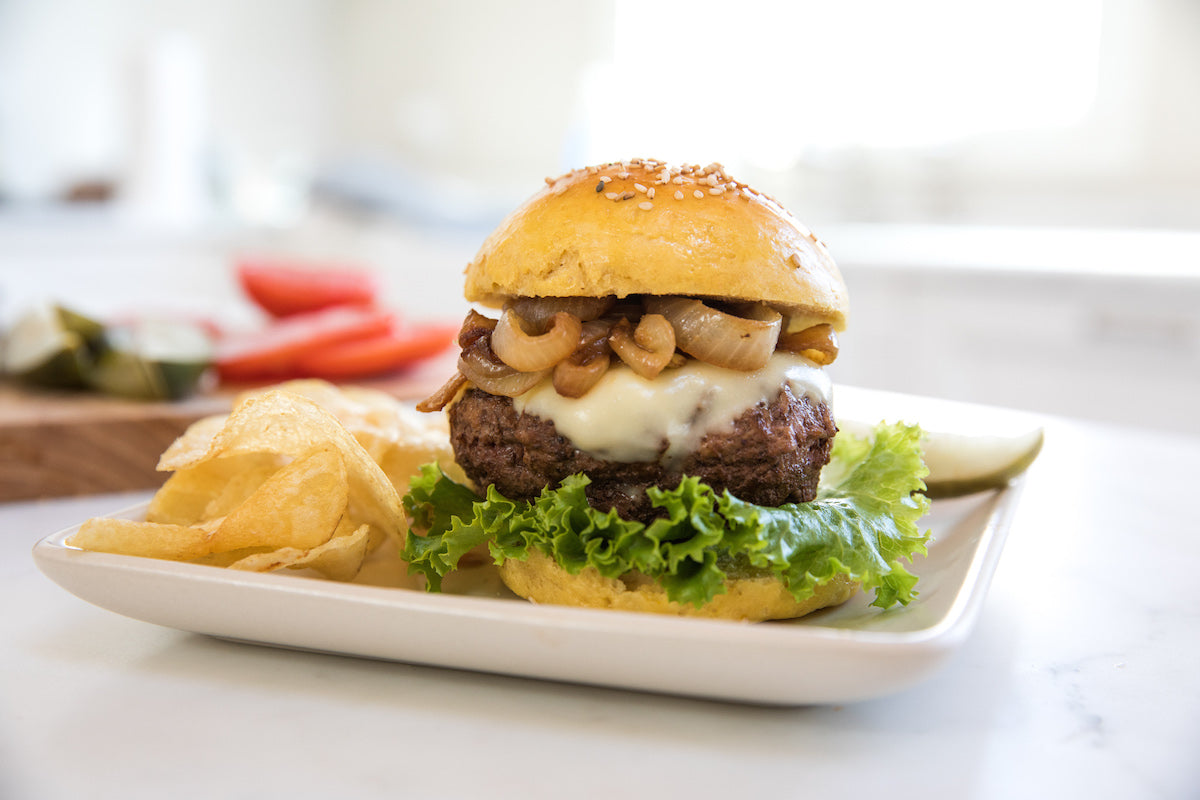 Einkorn hamburger bun sandwich with melted cheese, sautéed onions and lettuce on a white plate with potato chips and a pickle in a bright kitchen