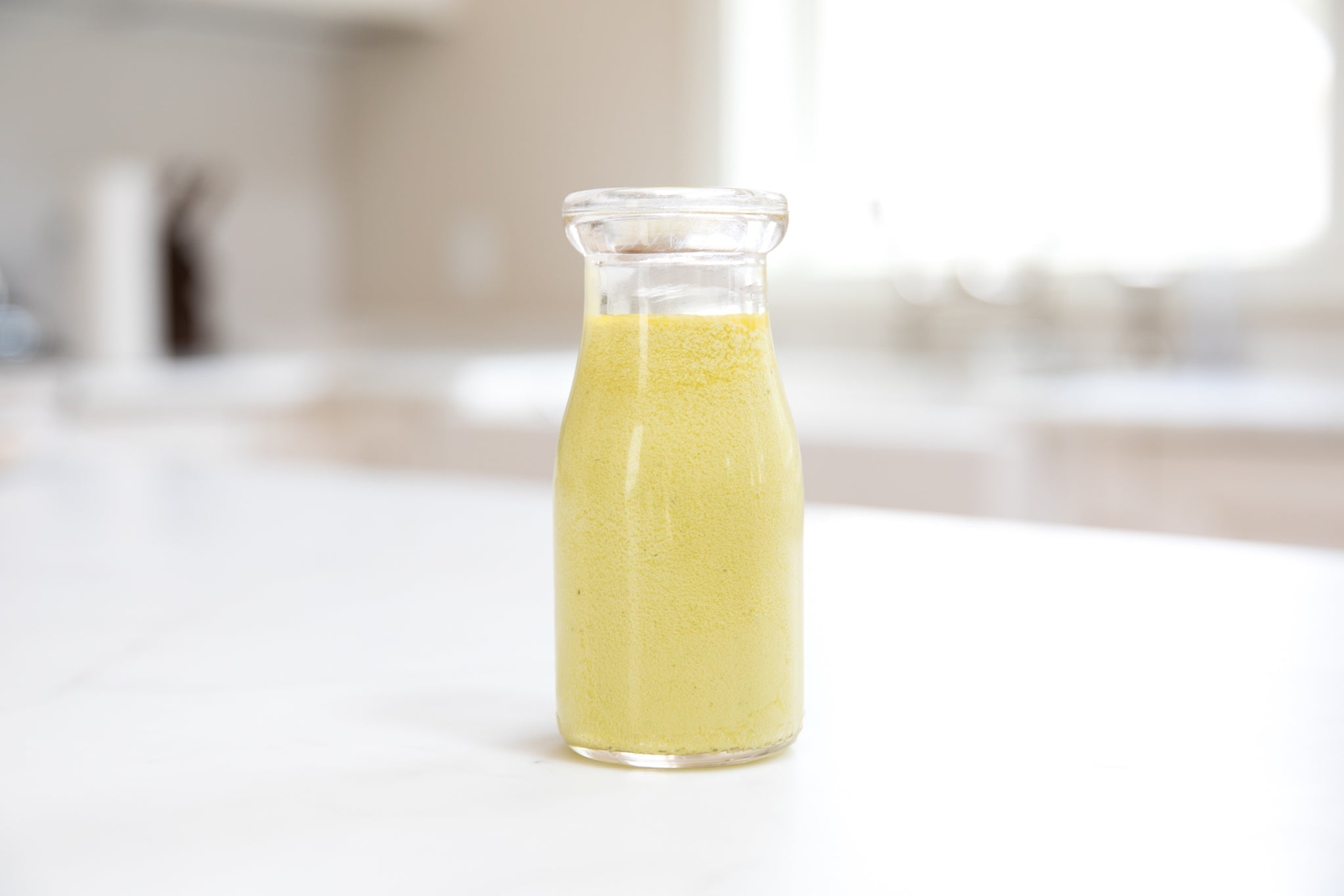 Close-up of small glass bottle of creamy herb and olive oil dressing showing smooth pale yellow emulsion on white countertop