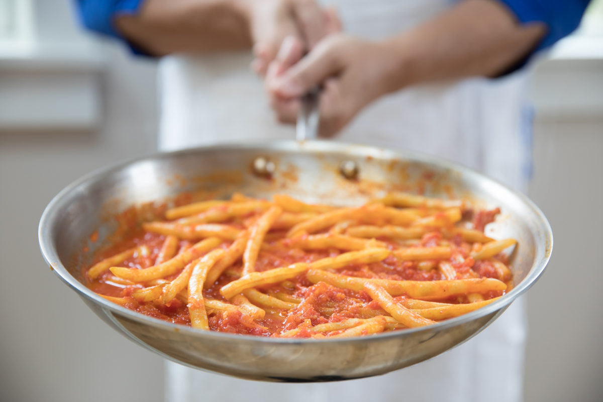Hands holding a stainless-steel skillet of tomato-braised yellow wax beans in a kitchen