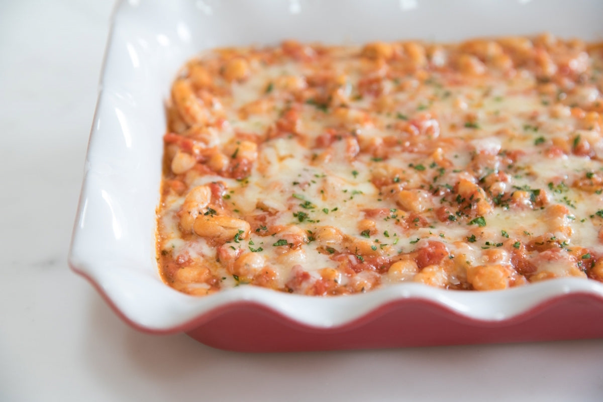 Close-up of baked cannellini beans in a red scalloped baking dish topped with melted cheese and chopped parsley on a marble countertop