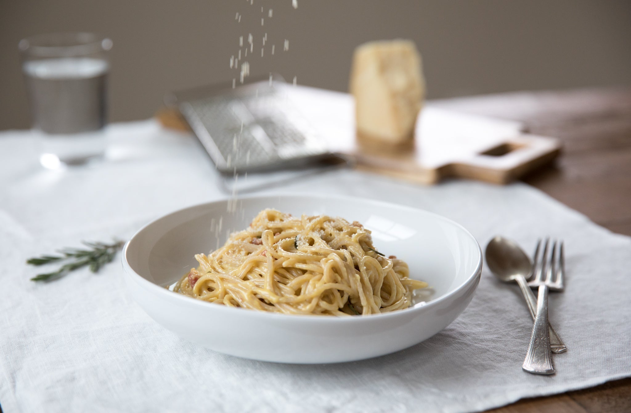 Spaghetti carbonara with pancetta being dusted with grated cheese in a white bowl on a linen napkin, utensils at right
