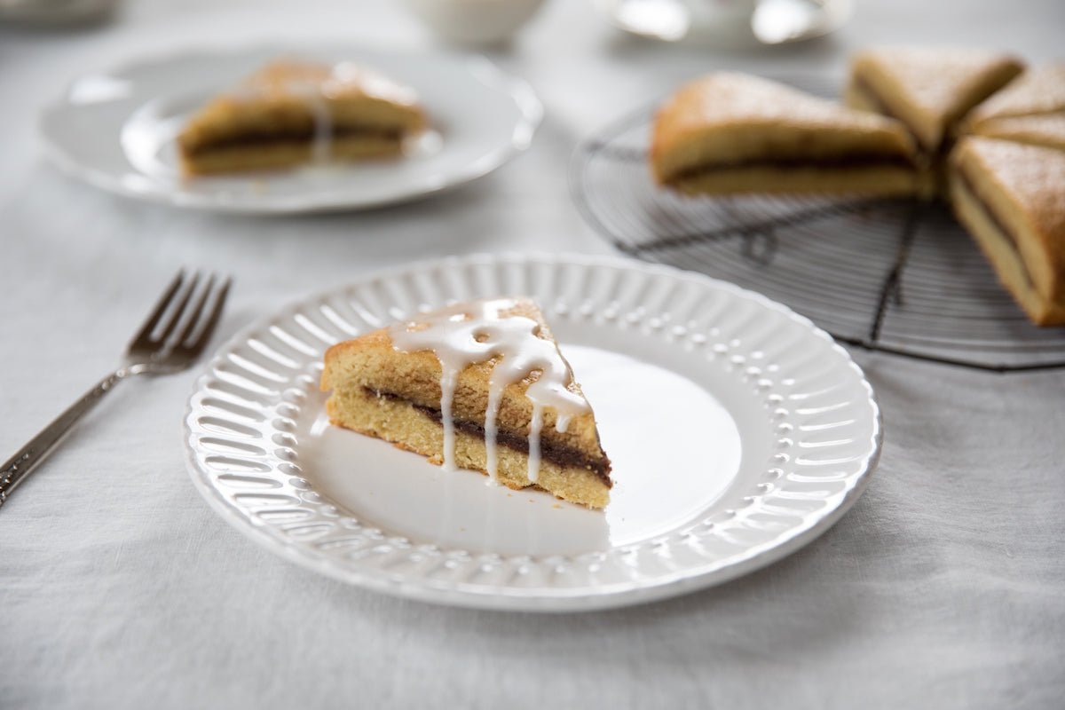 Slice of Einkorn cinnamon scone with icing drizzle on a white plate, fork at left and scones on a cooling rack on a linen tablecloth