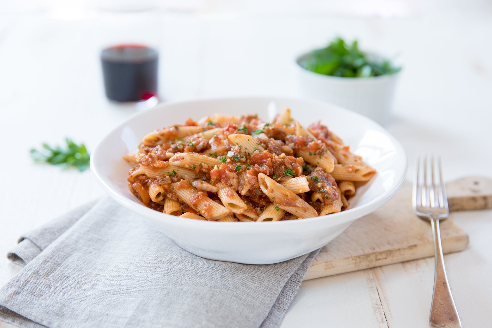 Bowl of penne with vegan eggplant and mushroom Bolognese on a white table with fork and glass of red wine