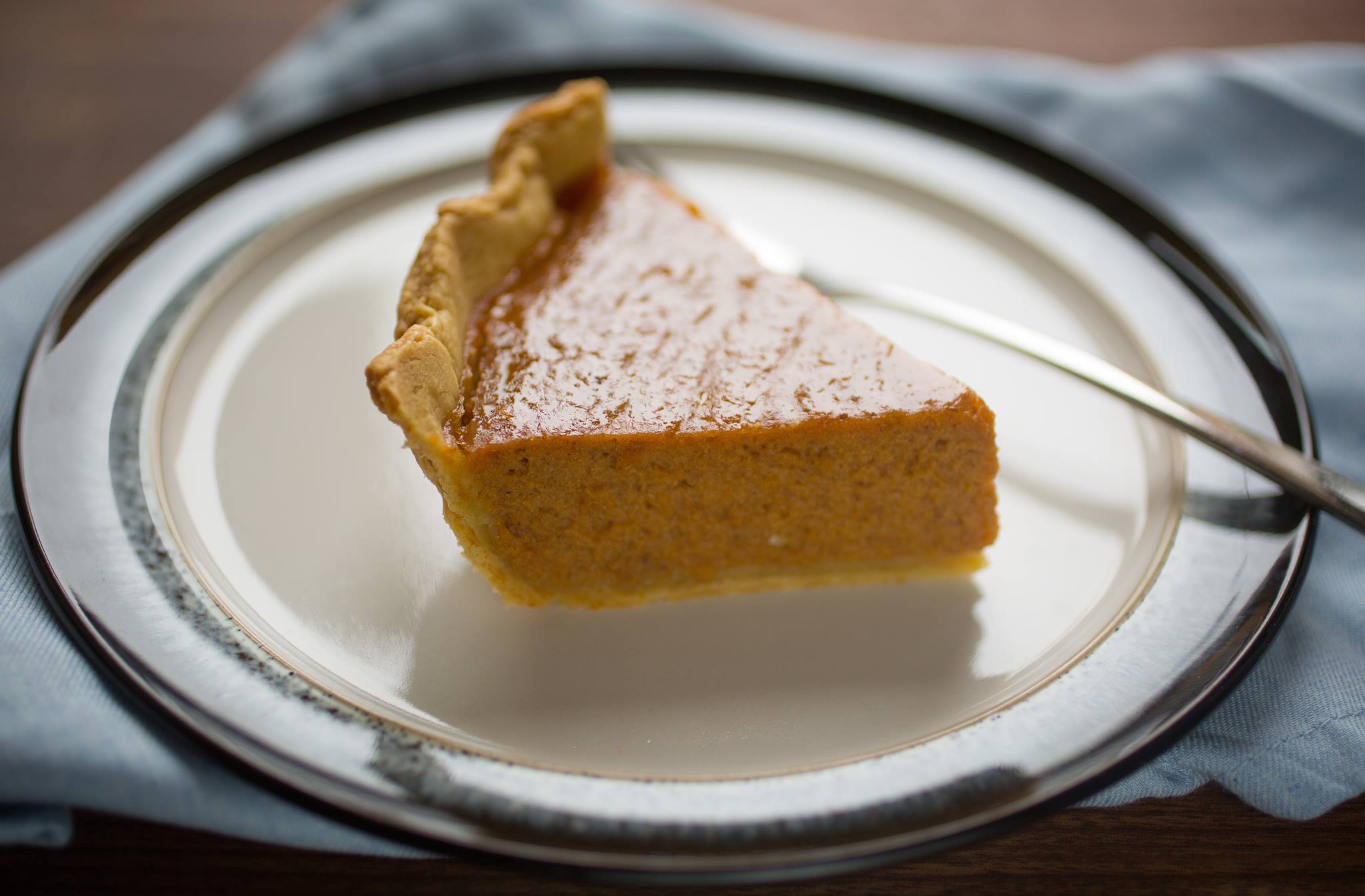 Slice of pumpkin pie with golden flaky crust on a white plate, fork to the side and a blue napkin underneath