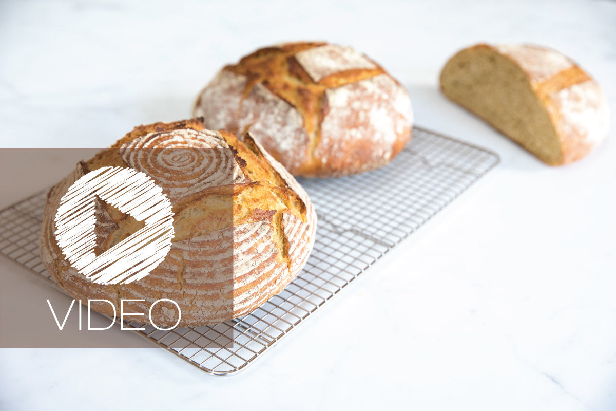 Two whole Einkorn sourdough boules and a halved loaf cooling on a wire rack on a light countertop with a VIDEO play overlay