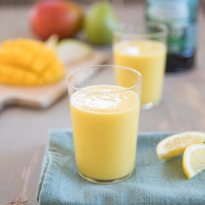 Glass of pear and mango smoothie made with olive oil, served on a cloth with sliced mango and lemon wedges blurred in the background