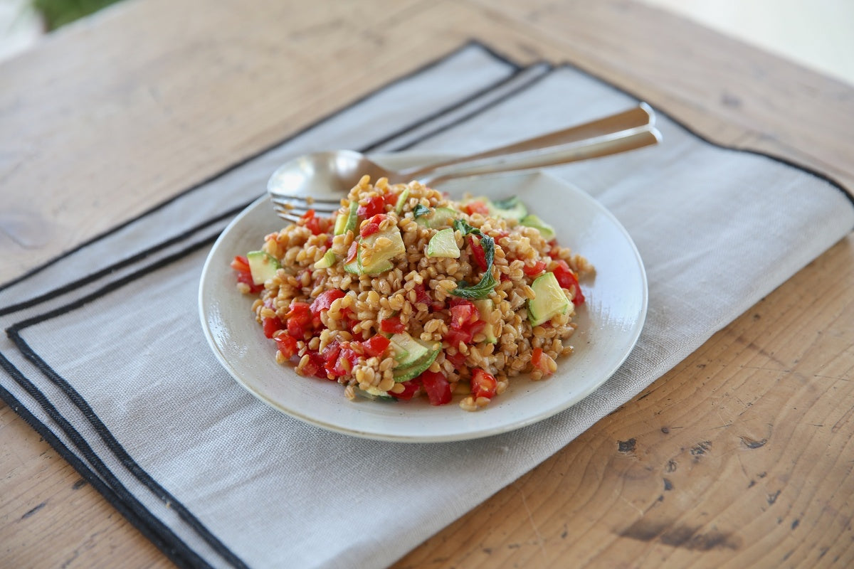 Ancient grain Einkorn wheat berry salad with tomatoes, avocado, and herbs served on a white plate on a linen napkin on a wooden table