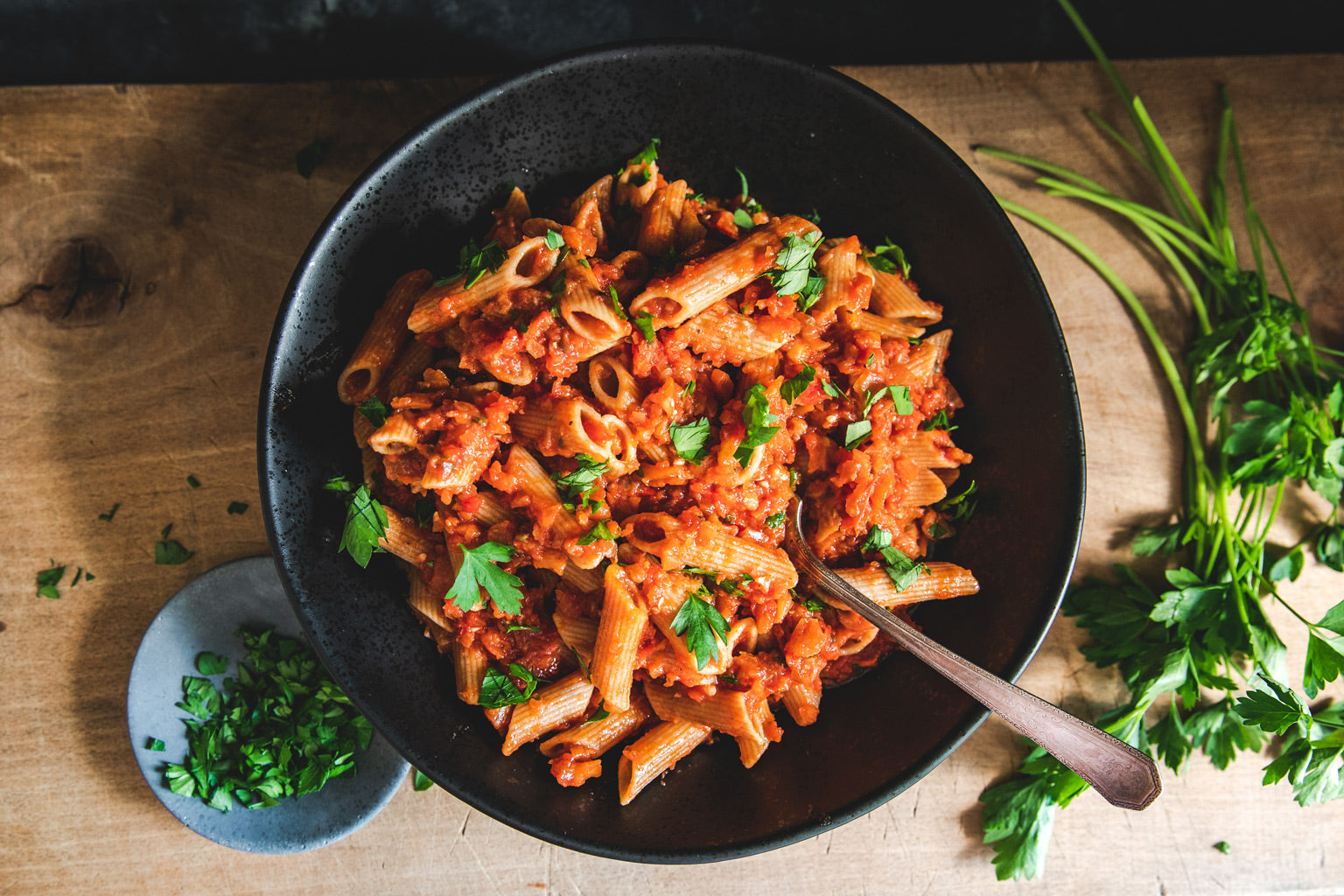 Bowl of penne pasta with eggplant bolognese, garnished with parsley, on a wooden table