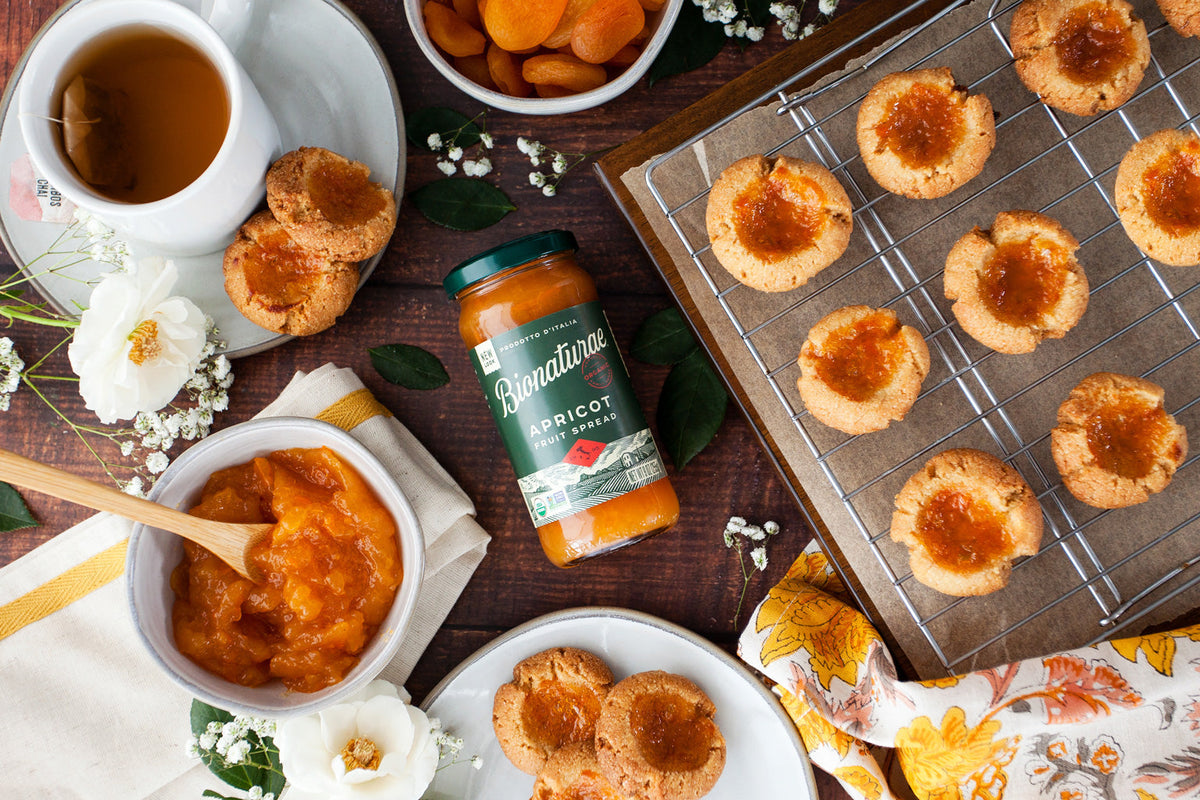 Jar of Bionaturae apricot fruit spread beside apricot thumbprint cookies on a cooling rack and plates in a rustic tabletop setting
