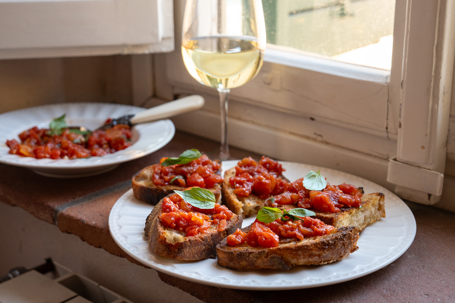 Tomato bruschetta on toasted bread served on a white plate by a sunlit windowsill with a glass of white wine