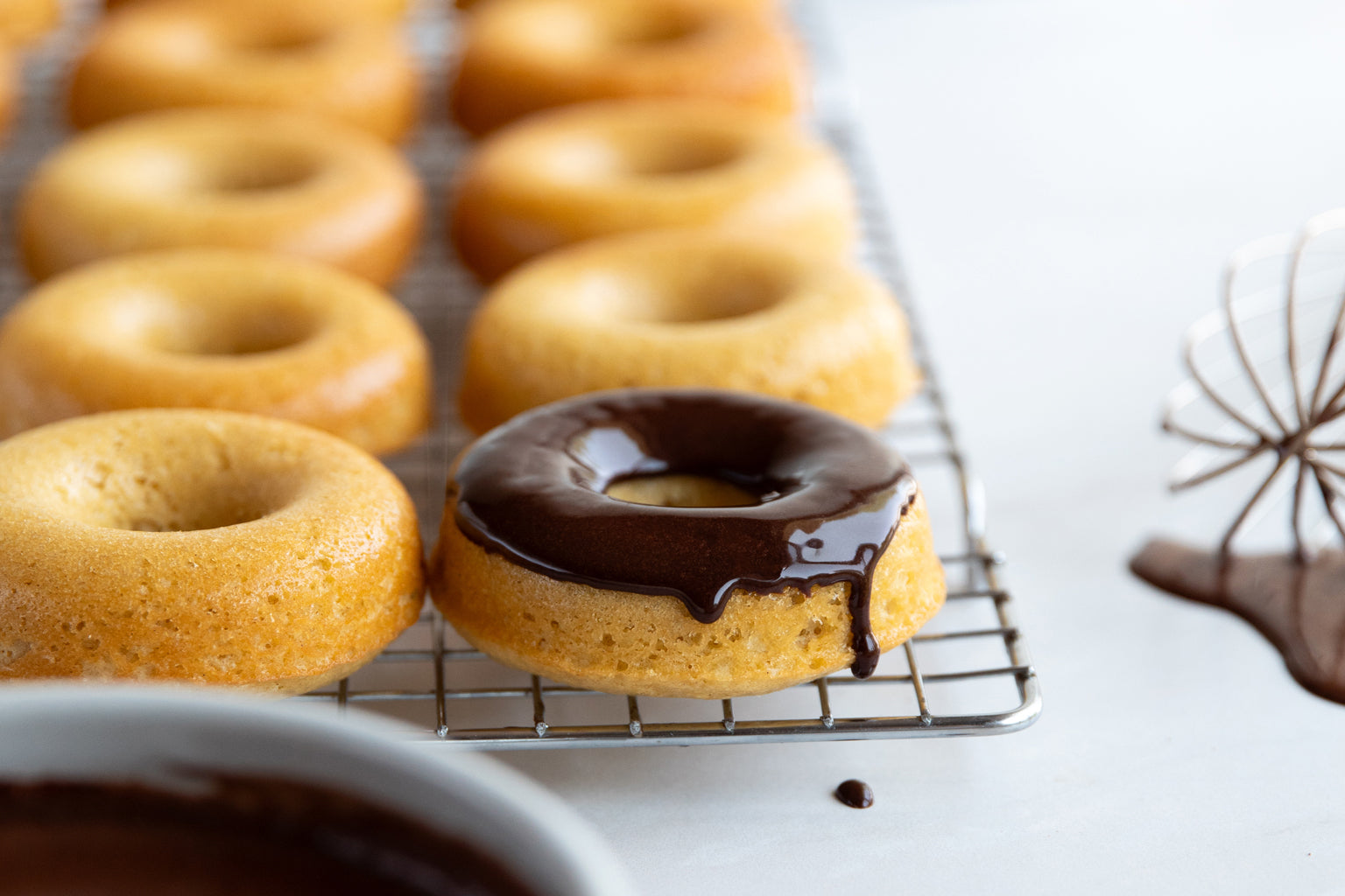 Rows of einkorn donuts on a cooling rack, one topped with dripping chocolate frosting