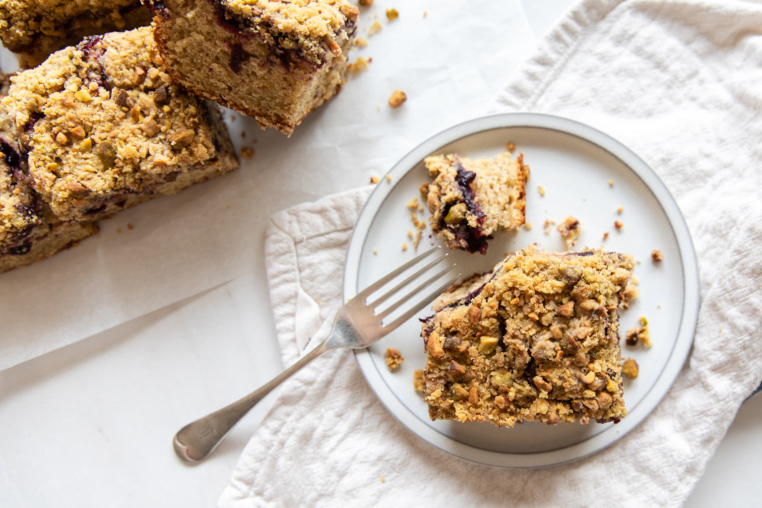 A slice of Einkorn Cherry Pistachio Coffee Cake on a plate with a piece cut off, next to rows of coffee cake slicesA slice of Einkorn Cherry Pistachio Coffee Cake on a plate