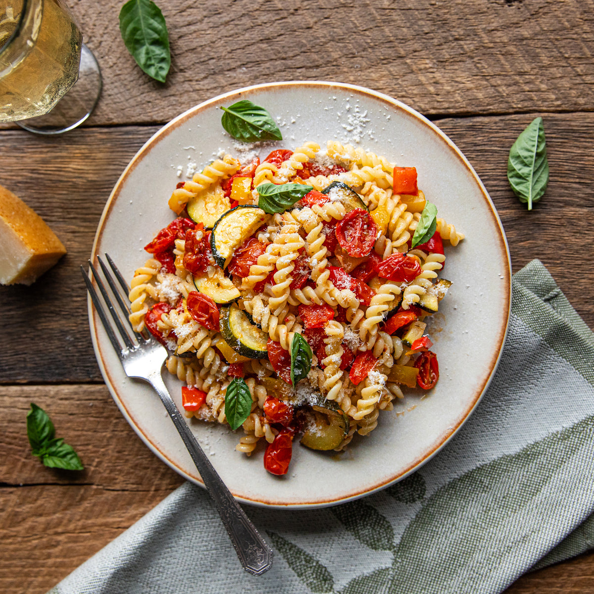 Plate of fusilli with oven-roasted tomatoes, zucchini, red pepper, basil, and grated Parmesan on a wooden table